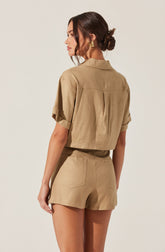A woman is wearing a beige, short-sleeved, cropped jacket with rolled cuffs and a collar, paired with matching high-waisted shorts that have visible back pockets.